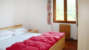 Una cama individual con una manta roja, cabecera de madera, mesita de noche con lámpara y ventana con cortinas florales.