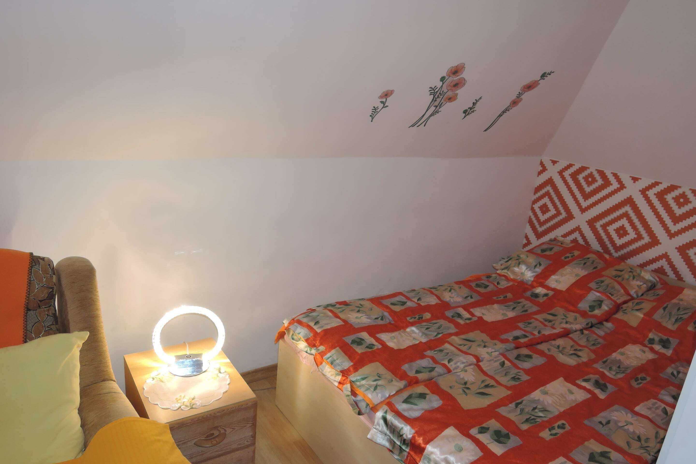 A bedroom with a bed, a bedside table with a lamp, a chair, and a wall with floral decorations.