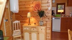 A wooden cabin interior with a wooden cabinet, a lamp, a plant, a chair, a small table, and a kitchen area with a dishwasher and a clock. - Holiday House in Sarbinowo Near Baltic Beach (Mielno)