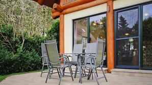An outdoor patio with a table and four chairs, surrounded by greenery and a building with large windows. - Ferienhaus Waldblick in Gohren-lebbin (Goehren-Lebbin)