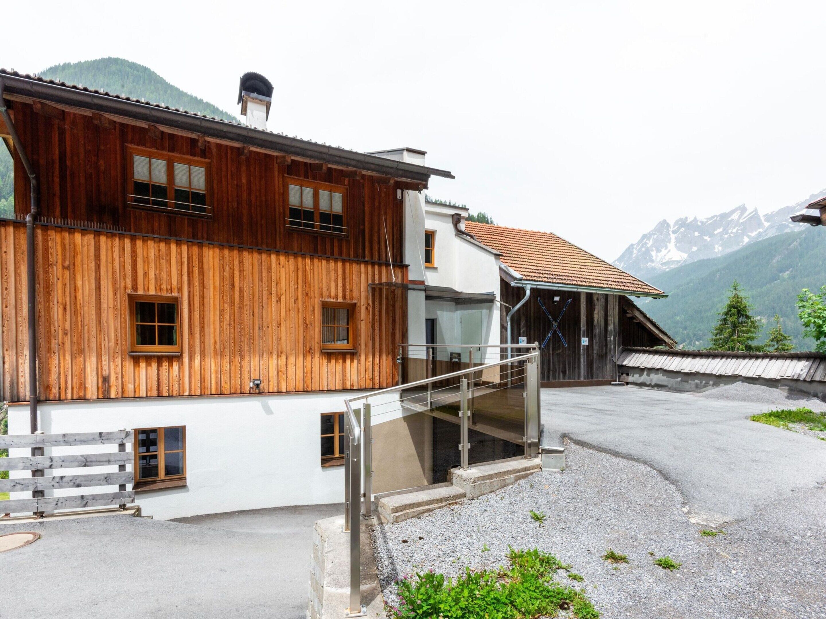 A wooden chalet with a white upper section, a gravel driveway, and a mountain backdrop.
