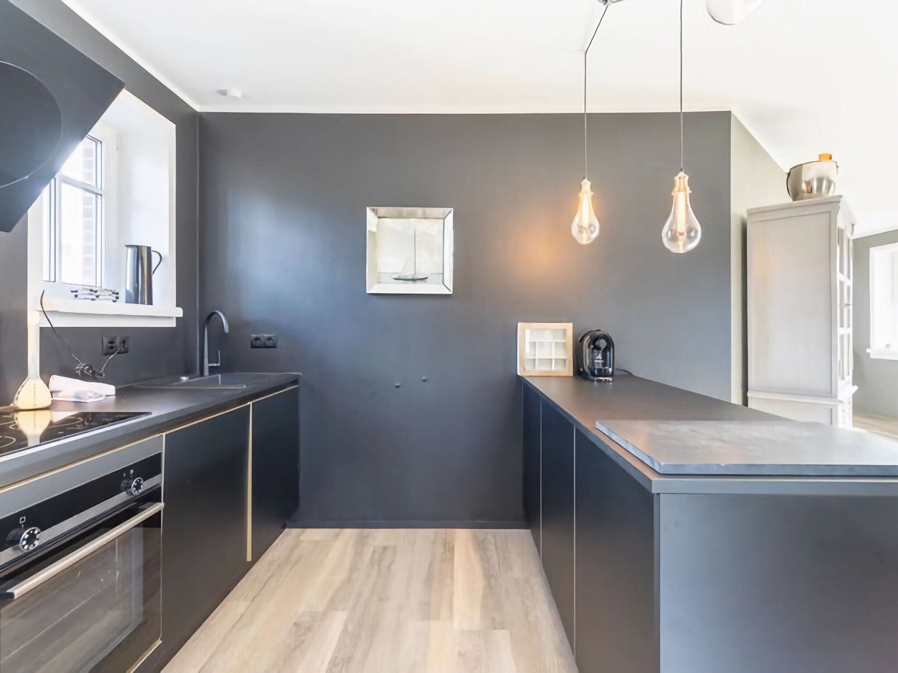 A modern kitchen with dark walls, a wooden floor, a built-in oven, and pendant lighting.