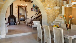 A spacious living area with a stone wall, a fireplace, wicker furniture, and a dining table with fruit. - Cozy Holiday Home With Pool and Whirlpool in Maheri (Apokoronas)