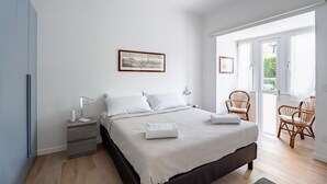 Un dormitorio con cama, mesita de noche, lámpara, cuadro enmarco, sillas de mimbre y una puerta que da a un balcón.