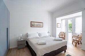 A bedroom with a bed, nightstand, lamp, framed picture, wicker chairs, and a door leading to a balcony.