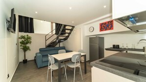 A compact living space with a dining area, a kitchenette, a staircase, and a flat-screen TV mounted on the wall.