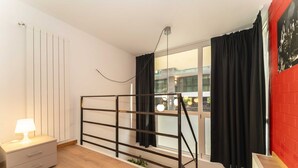 A room with a bed, a nightstand, a window with curtains, a radiator, and a red brick wall.