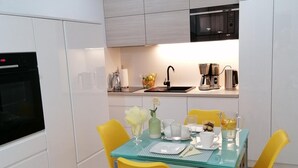 A compact kitchen with white cabinets, a black oven, a stainless steel coffee maker, and a yellow dining table set with glassware and a vase. - Apartment Near Baltic Sea With Balcony View (Rewal)