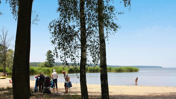 Egy tóparti helyszín, ahol emberek vannak a víz közelében, fák és egy homokos strand láthatók.