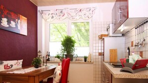 Egy praktikus, kis méretű konyha, étkezősarokkal, egy ablakkal, melyből zöld tájra nyílik kilátás, és egy piros székkel.