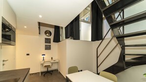 Une cuisine compacte dotée d’un four encastré, un coin repas avec une table et des chaises, un petit bureau avec une lampe et un escalier avec une rampe noire.