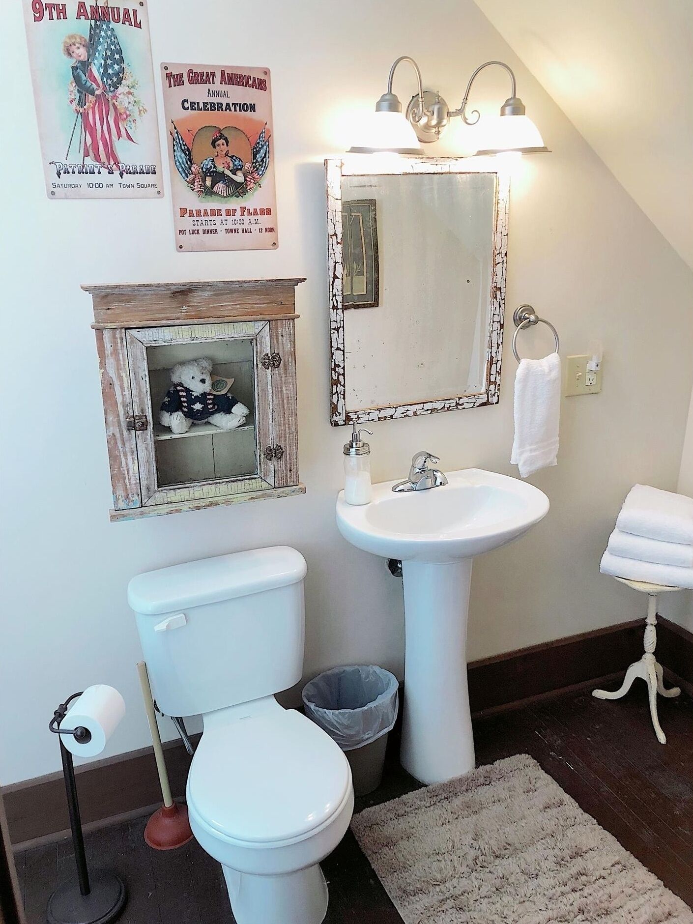 A bathroom with a white toilet, a white sink, and a mirror above the sink. There is a poster on the wall and a small cabinet with a teddy bear.