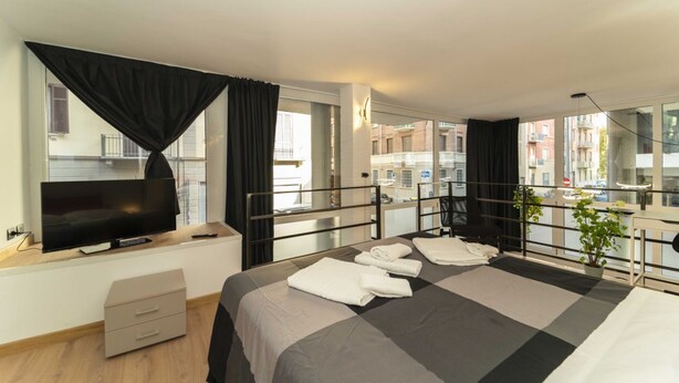 A hotel room with a bed, a TV, a desk, and a balcony with a view of buildings. - GuestHost - Galaxy Lofts - Appt Uranus x4 (Torino)