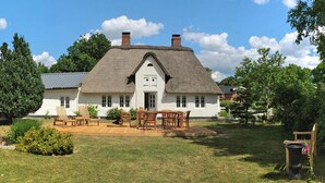 A white house with a thatched roof, a front porch with wooden chairs and a table, and a well-maintained lawn. - Reetgedecktes Ferienhaus in Humptrup (Humptrup)