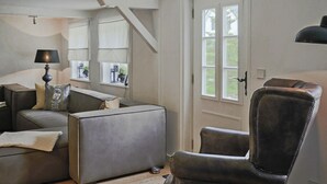 A modern living room with a sofa, armchair, and a lamp. There is a window with curtains and a door with a handle. - Reetgedecktes Ferienhaus in Humptrup (Humptrup)
