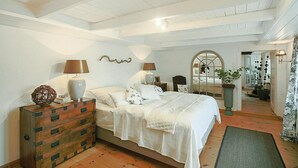 A bedroom with a wooden dresser, two bedside tables with lamps, a large bed with white bedding, a mirror, and a potted plant. - Reetgedecktes Ferienhaus in Humptrup (Humptrup)