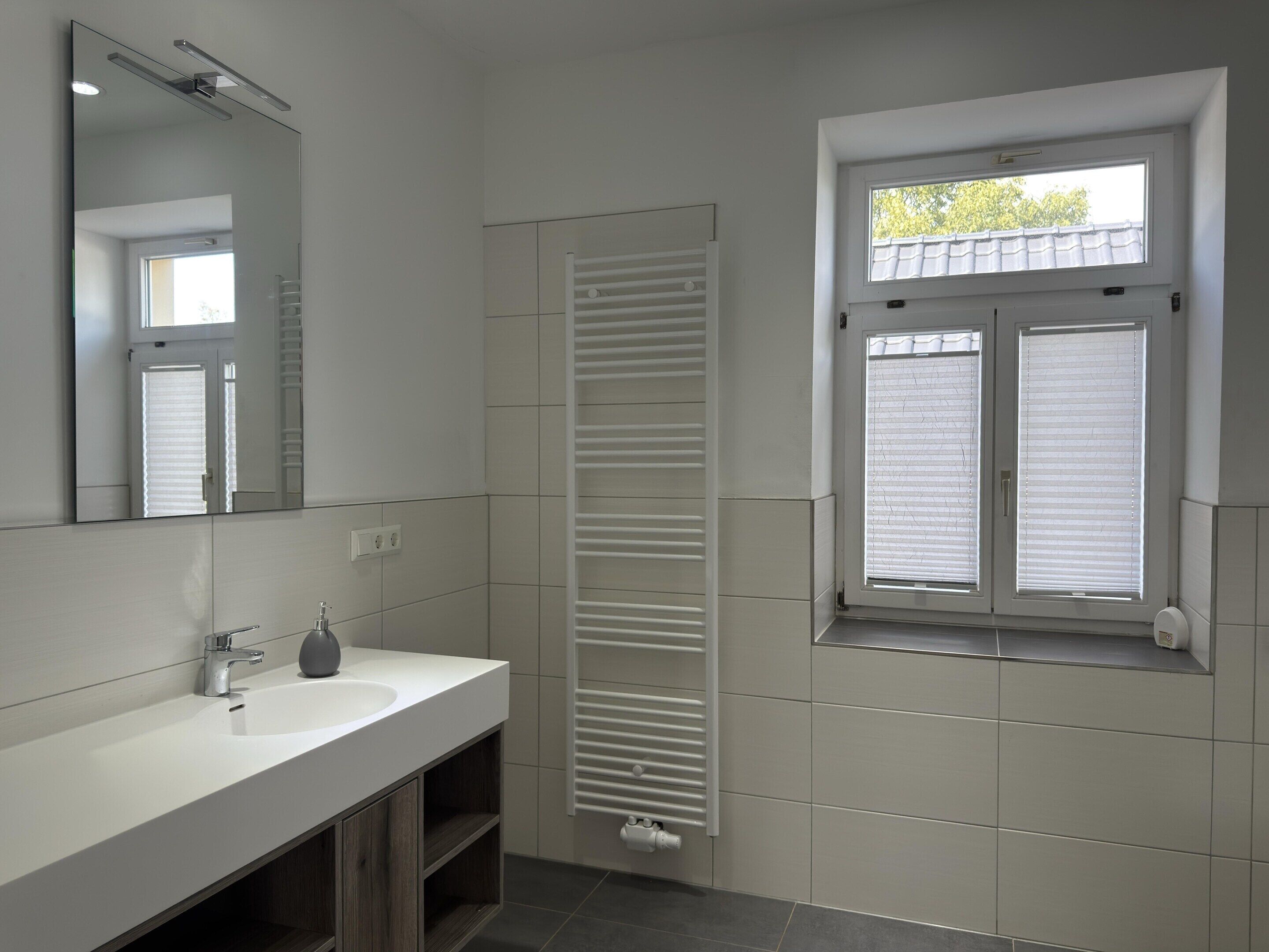 Een moderne badkamer met een witte wastafel, spiegel en raam met zonwering.