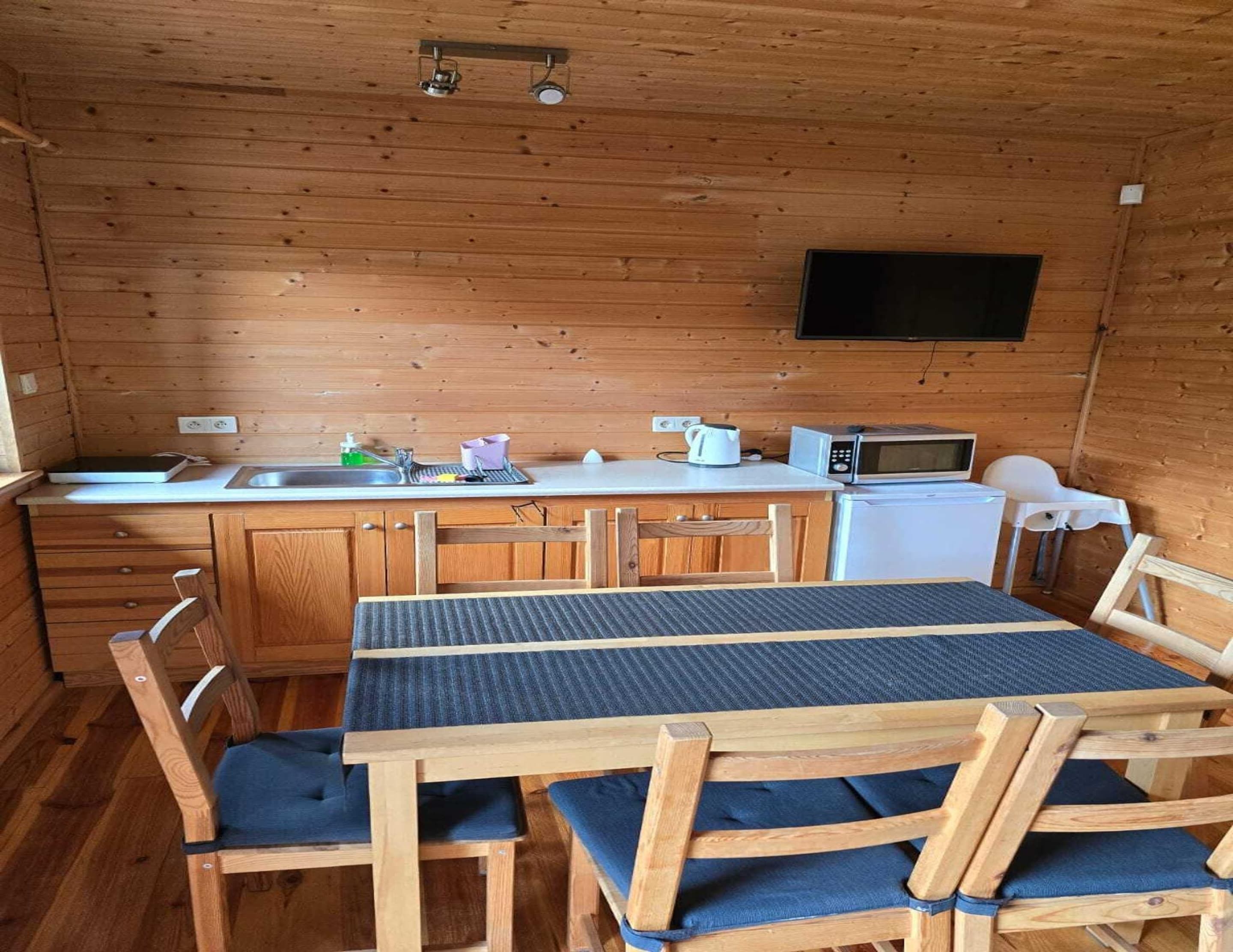 A compact kitchen with wooden cabinets, a white countertop, a microwave, a sink, and a small refrigerator. There is a table with chairs and a flat-screen TV mounted on the wall.