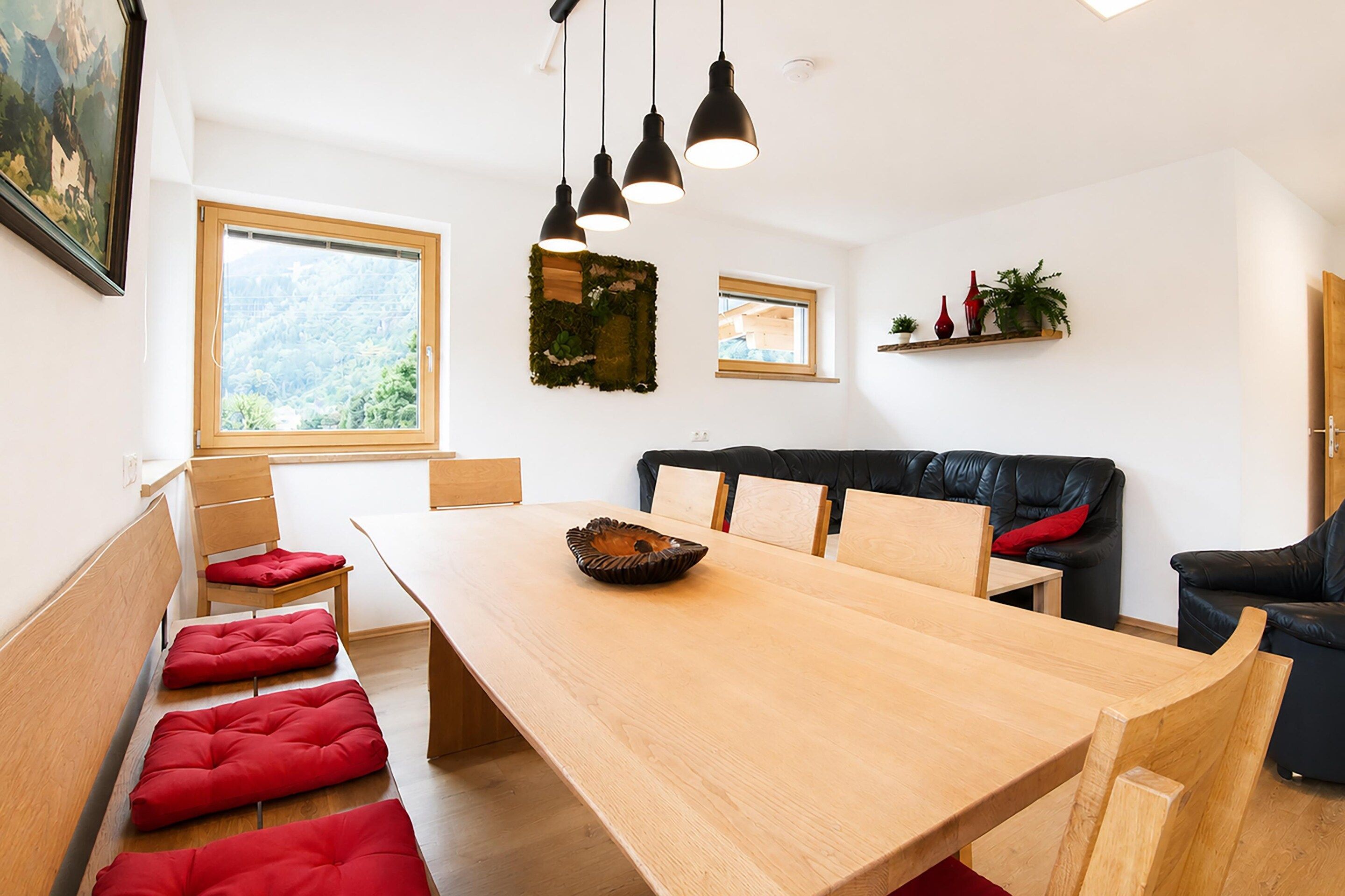 A modern interior with a wooden dining table, red cushioned benches, a black leather sofa, and pendant lighting.