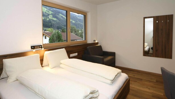 Una cama doble con ropa de cama blanca, cabecera de madera, ventana con vistas a un pueblo, suelo de madera y un espejo en la pared.