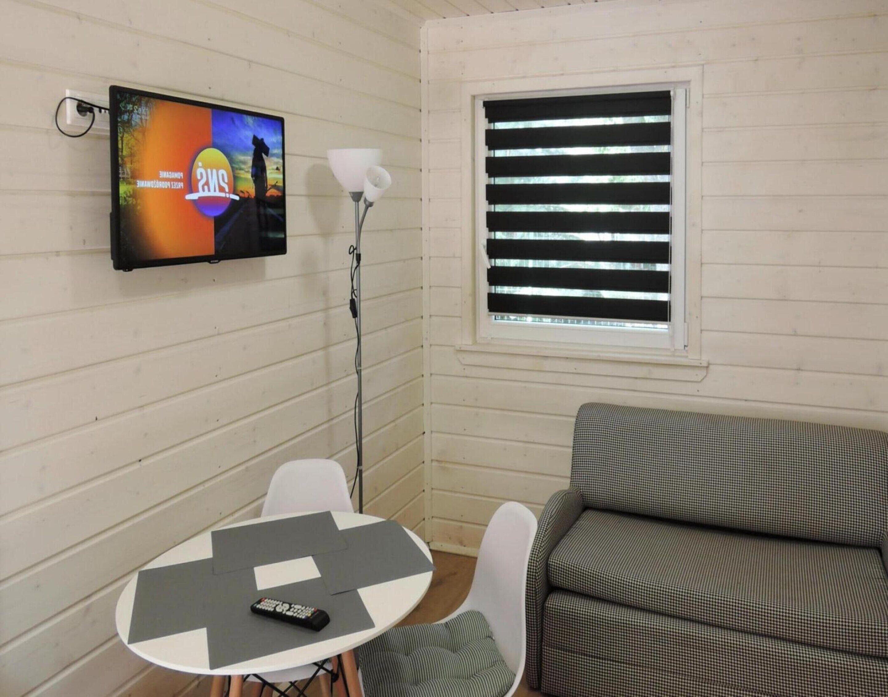 A room with a TV mounted on the wall, a table with a remote control, a lamp, and a window with blinds.