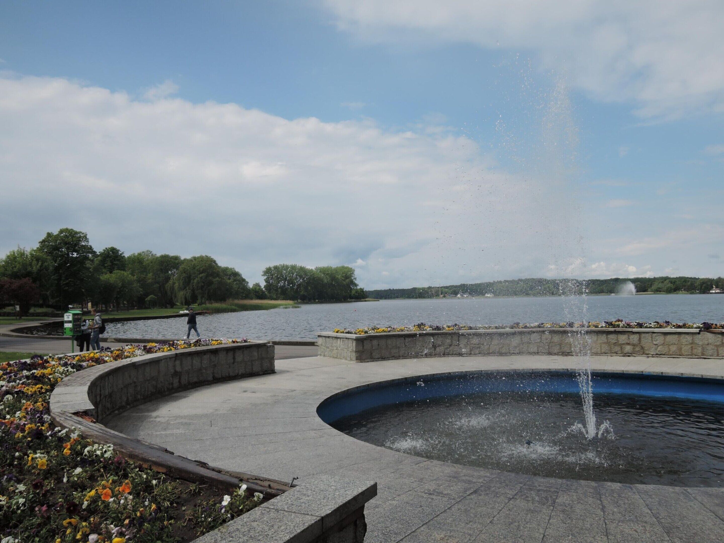 A circular fountain with water jets, surrounded by a paved walkway and flower beds.