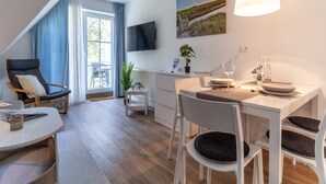 A modern, well-lit living area with a dining table, chairs, a sofa, a TV mounted on the wall, and a large window with curtains. - Ferienwohnung in St. Peter-ording (St. Peter-Ording)