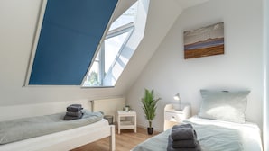 A modern bedroom with a sloped ceiling, two beds, a small bedside table, a nightstand, a lamp, a potted plant, and a painting on the wall. - Ferienwohnung in St. Peter-ording (St. Peter-Ording)