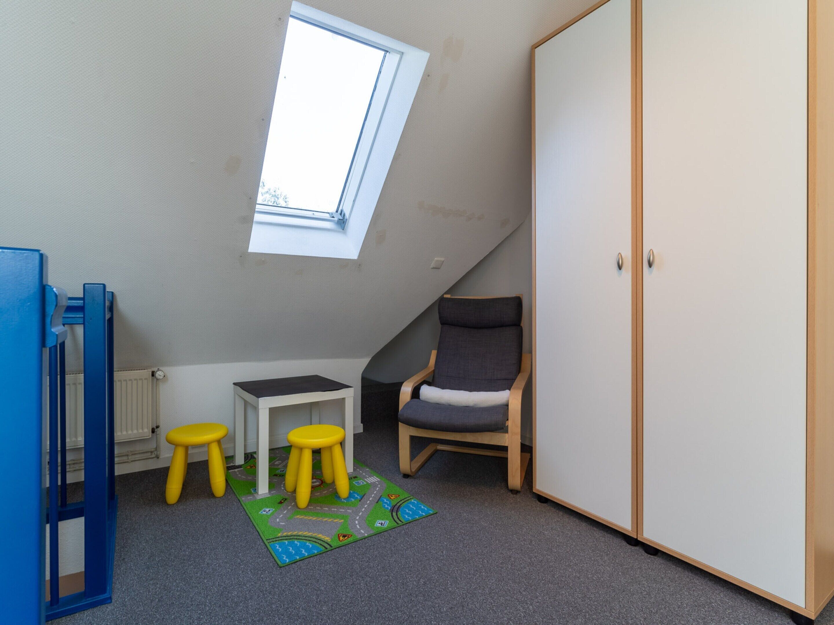 Una habitación con techo inclinado, tragaluz, una silla, armario y una pequeña mesa con taburetes.