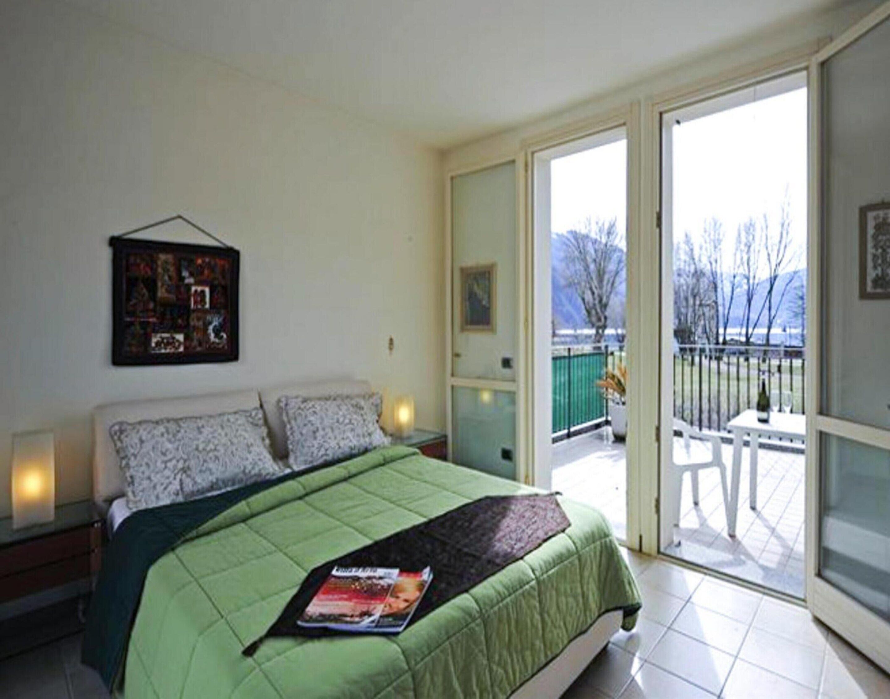 A bedroom with a bed, a nightstand, a wall-mounted artwork, and a sliding glass door leading to a balcony with a view of trees and mountains.