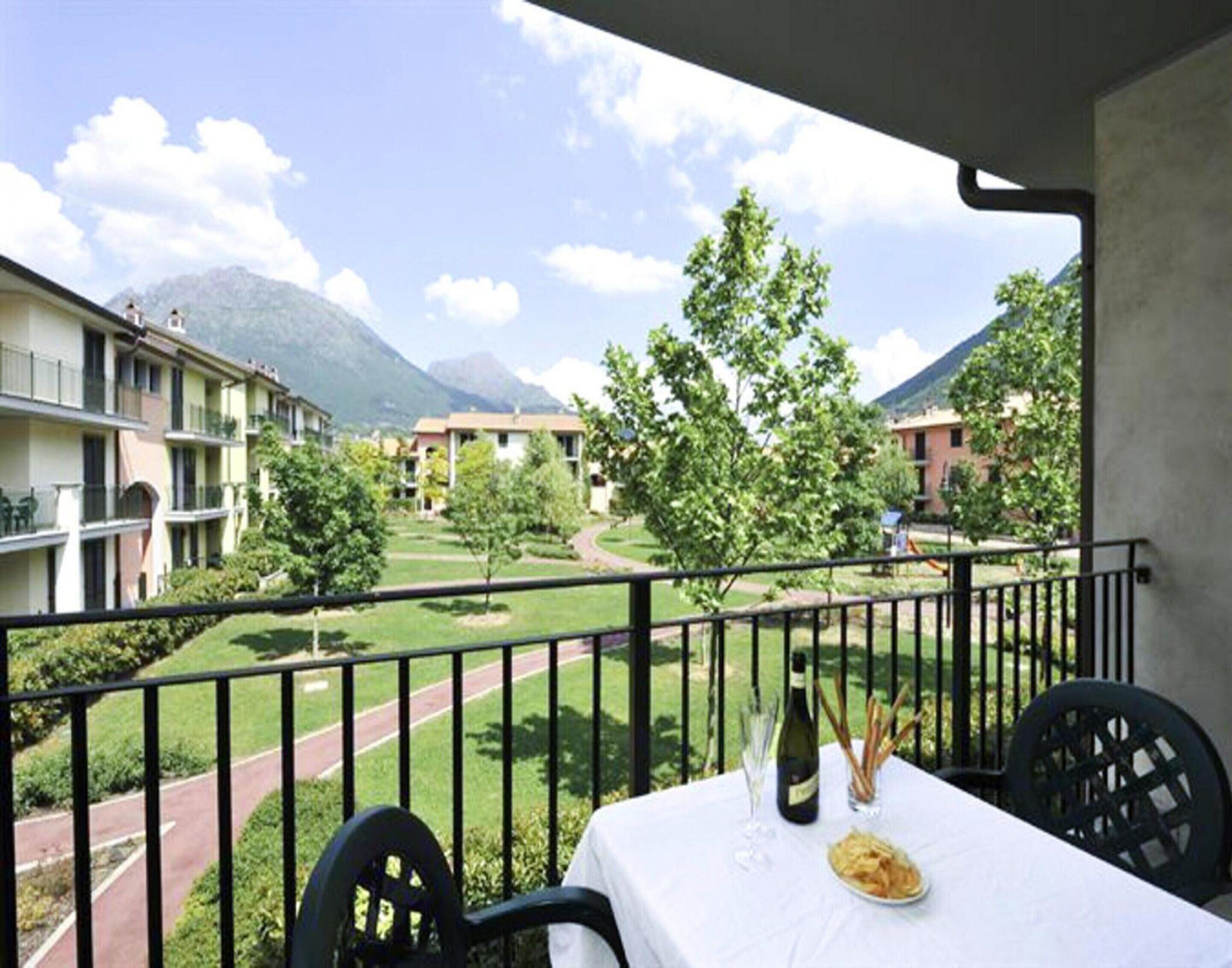 A balcony with a table set for dining, overlooking a courtyard with trees and a mountain in the background.