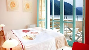 Ein Schlafzimmer mit einem Bett, einem Nachttisch, einer Lampe, einem roten Sessel und Blick auf einen See und Berge durch eine offene Tür.