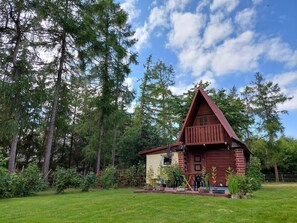 En stuga byggd av trä med ett triangulärt tak, omgiven av en välskött trädgård och höga träd.