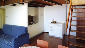 A compact kitchen with white cabinets, a stove, and a small countertop. There is a blue sofa and a wooden table with chairs in the living area. A wooden ladder is visible in the corner. - Residence Pratone, Garda (Garda)