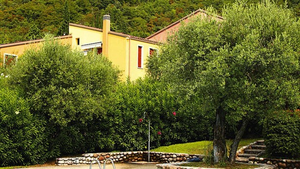 A swimming pool with a stone border, a yellow building with a red window, and a mountainous backdrop. - Residence Pratone, Garda (Garda)