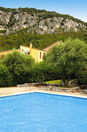 A swimming pool with a stone border, a yellow building with a red window, and a mountainous backdrop. - Residence Pratone, Garda (Garda)
