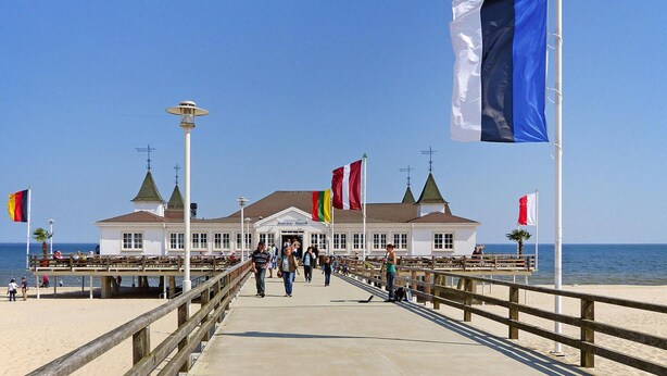 A pier leading to a building with multiple flags, a beach, and the sea in the background. - Doppelhaushalfte Dock No. 7.0 in Zirchow (Zirchow)