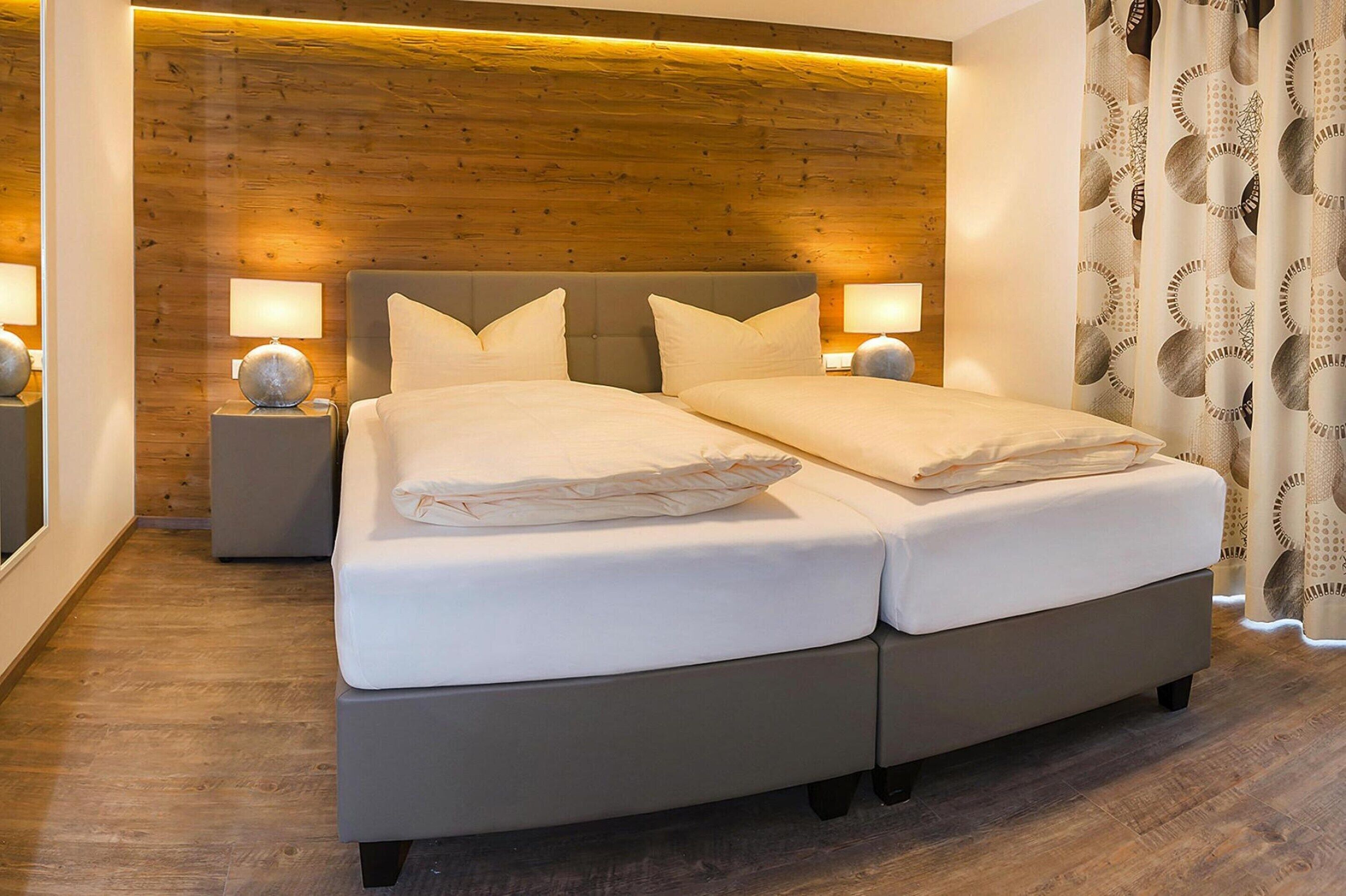 Un lit double avec une tête de lit grise et du linge de lit blanc, deux tables de chevet avec des lampes et un panneau mural en bois.