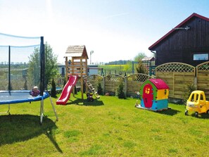 Un patio trasero cercado con un trampolín, una casita de juegos de madera, un tobogán y un pequeño coche de juguete amarillo.