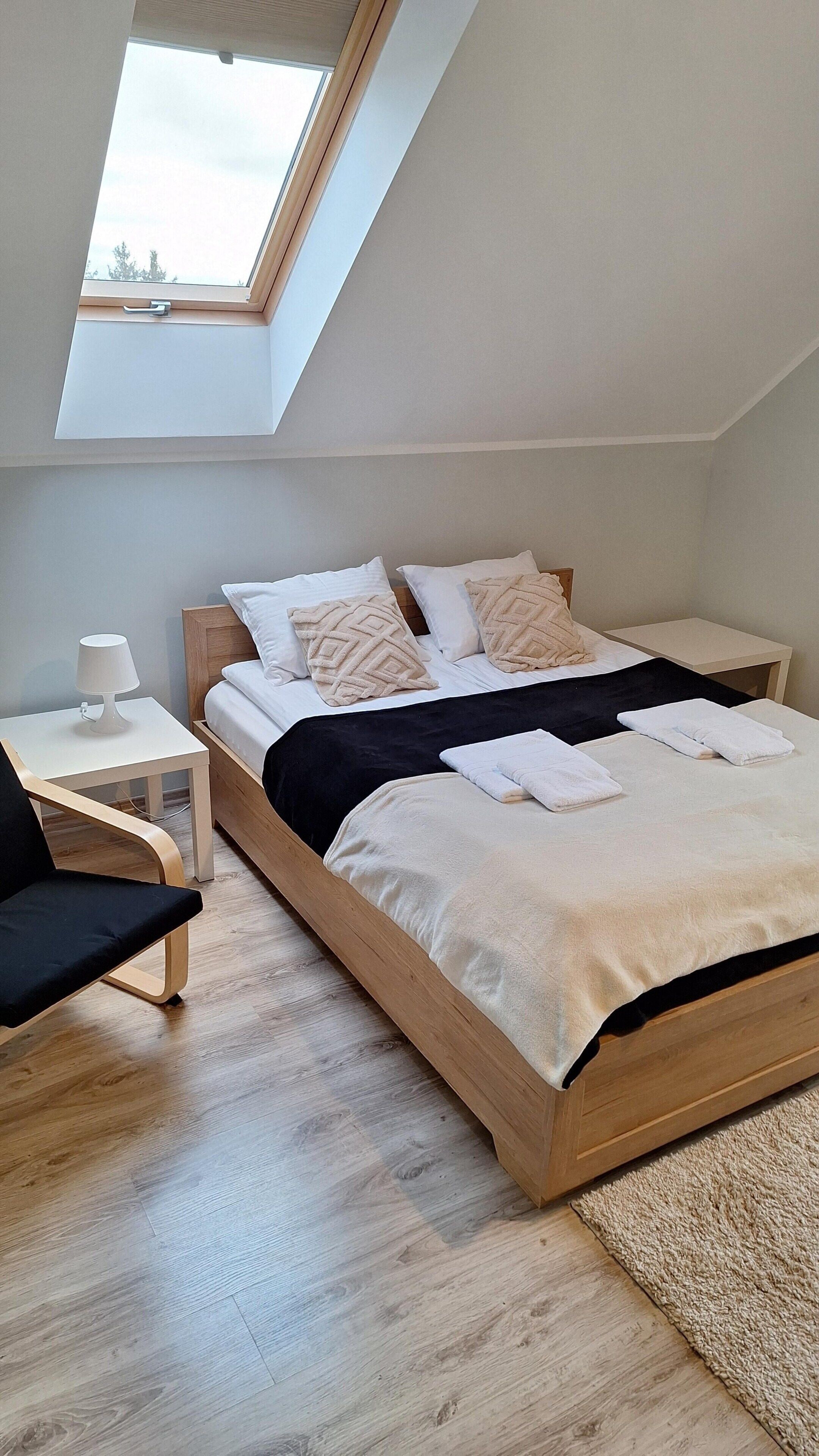 Un dormitorio moderno con un tragaluz, una cama con ropa de cama blanca, una mesita de noche con una lámpara y un suelo de madera.