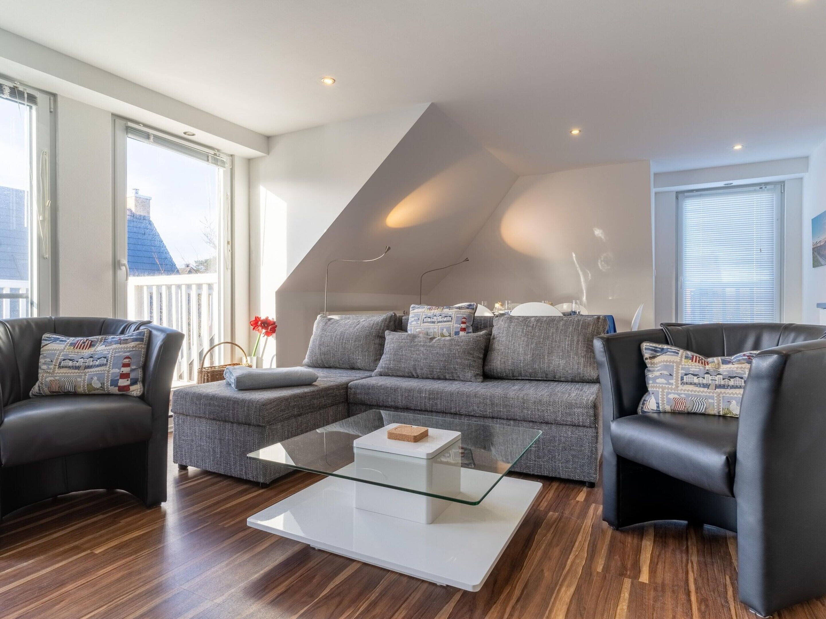 Uma sala de estar moderna com um sofá seccional cinza, duas poltronas pretas, uma mesa de centro de vidro e uma mesinha lateral branca.