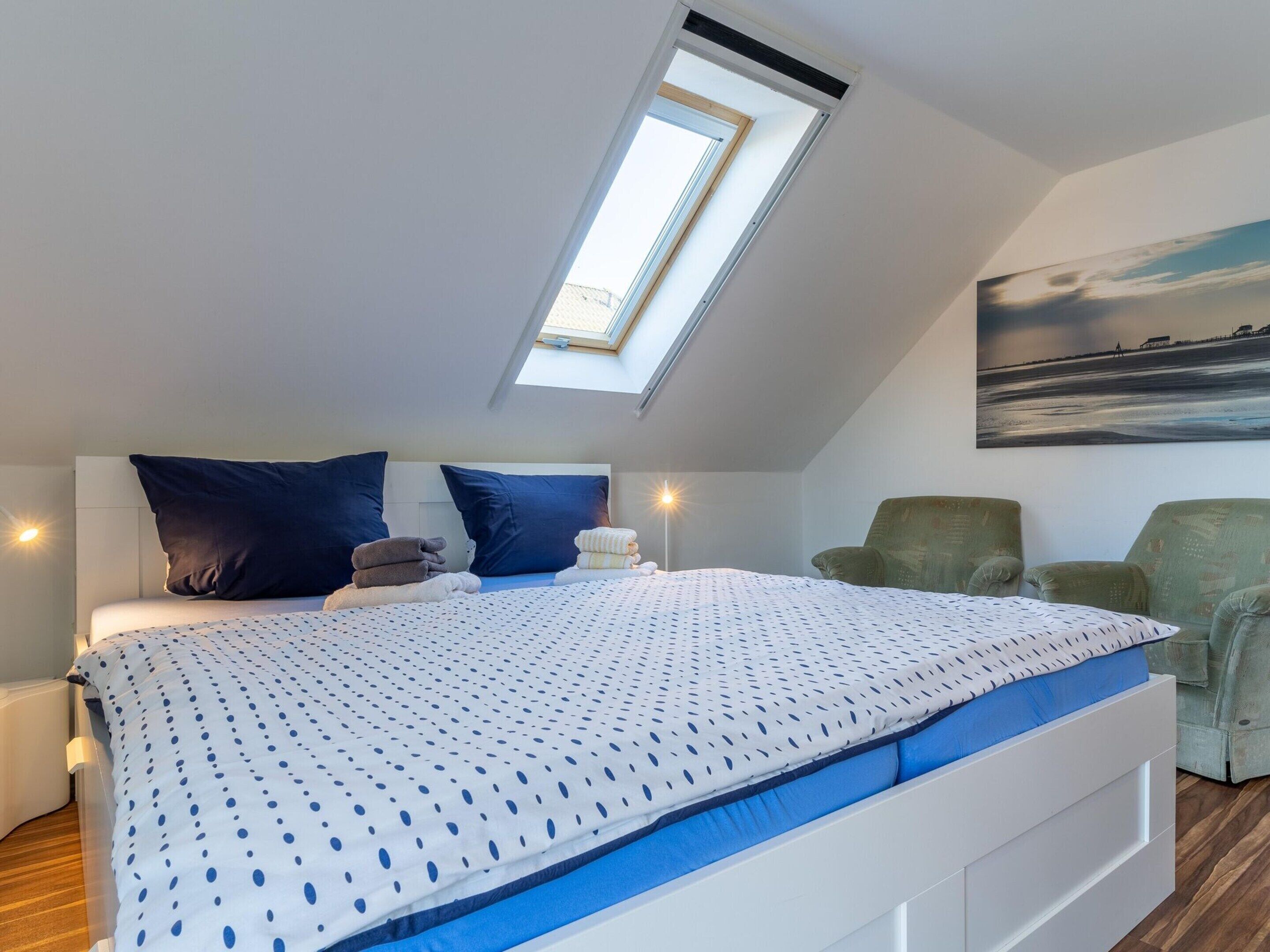 Une chambre avec un puits de lumière, un lit avec une literie bleue, deux oreillers, une table de chevet et deux chaises.