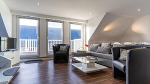 A modern living room with a sofa, armchairs, a coffee table, and a TV. Large windows offer a view of the outside. - Wohnung in St. Peter Ording (St. Peter-Ording)