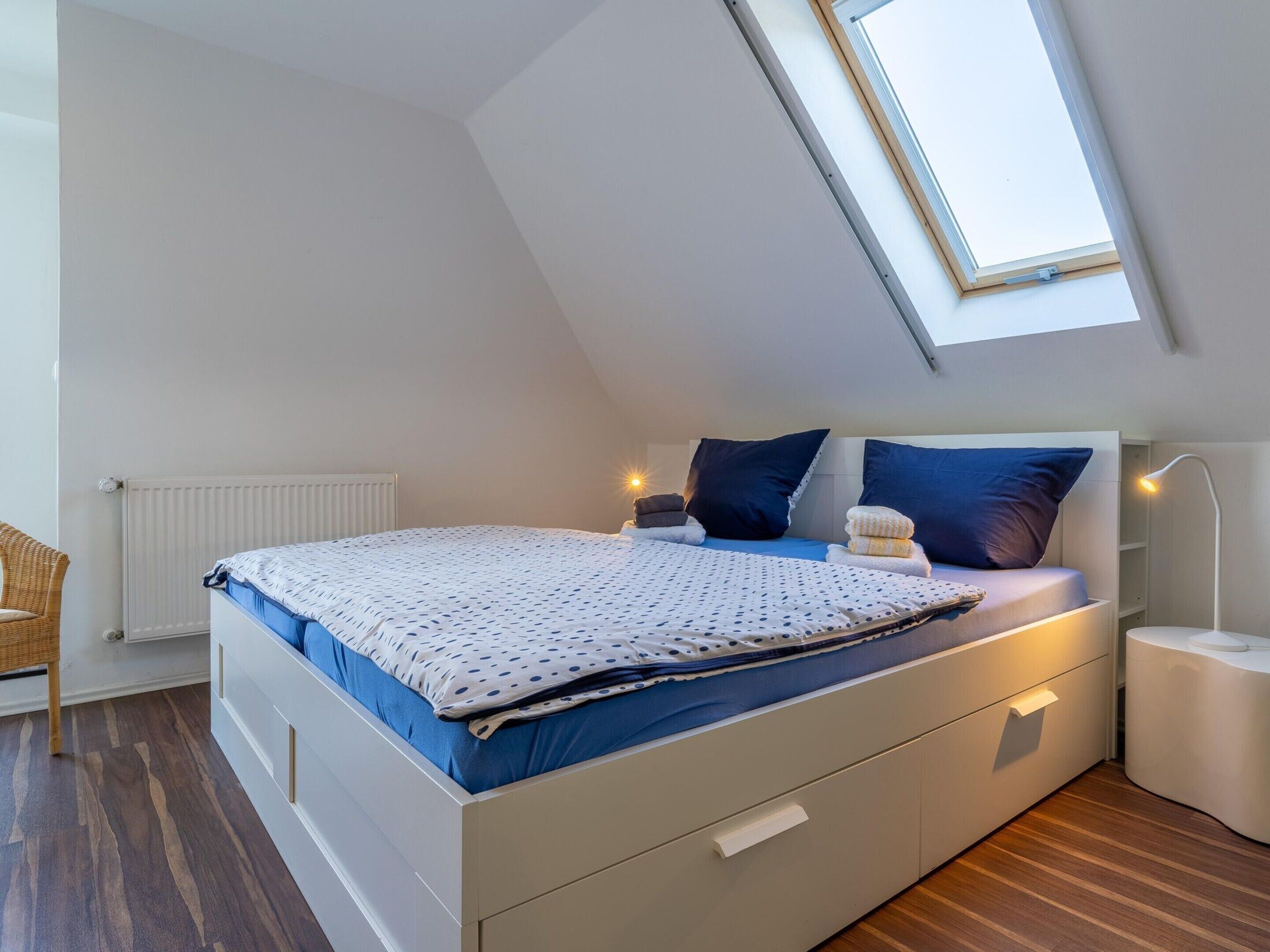 Ein modernes Schlafzimmer mit schrägem Dach, einem Bett mit gepunktetem Überwurf, zwei Kissen, einem Nachttisch mit Lampe und einem Heizkörper.