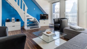 A modern living room with a blue accent wall, a white staircase, a flat-screen TV, a glass coffee table, and a balcony with a view. - Wohnung in St. Peter Ording (St. Peter-Ording)