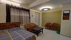 Une chambre à coucher avec un lit, une petite table, deux chaises en osier, une poubelle verte et une porte.