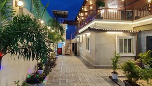Une cour intérieure avec un escalier menant à un balcon du deuxième étage, entourée de plantes en pot et illuminée par des guirlandes lumineuses.