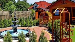 Une cabane en bois avec un toit rouge, une structure de jeu en bois et une fontaine dans la cour.