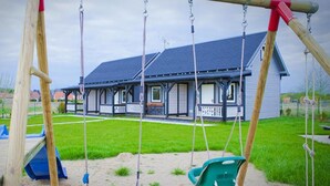 A playground with swings and a slide, a house with a porch, and a grassy yard. - Holiday Home in Rewal Near Baltic Beach (Rewal)
