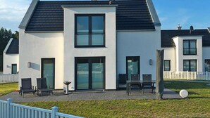 Une maison moderne à deux étages, dotée d’un toit plat, de grandes fenêtres et d’un patio aménagé avec du mobilier d’extérieur.